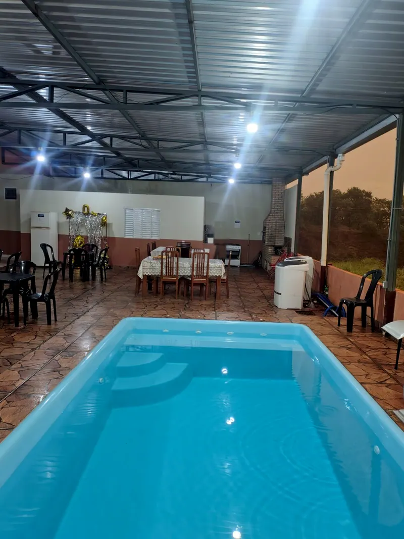 Piscina azul em primeiro plano, com a área gourmet ao fundo: mesas, churrasqueira de tijolinho e freezers