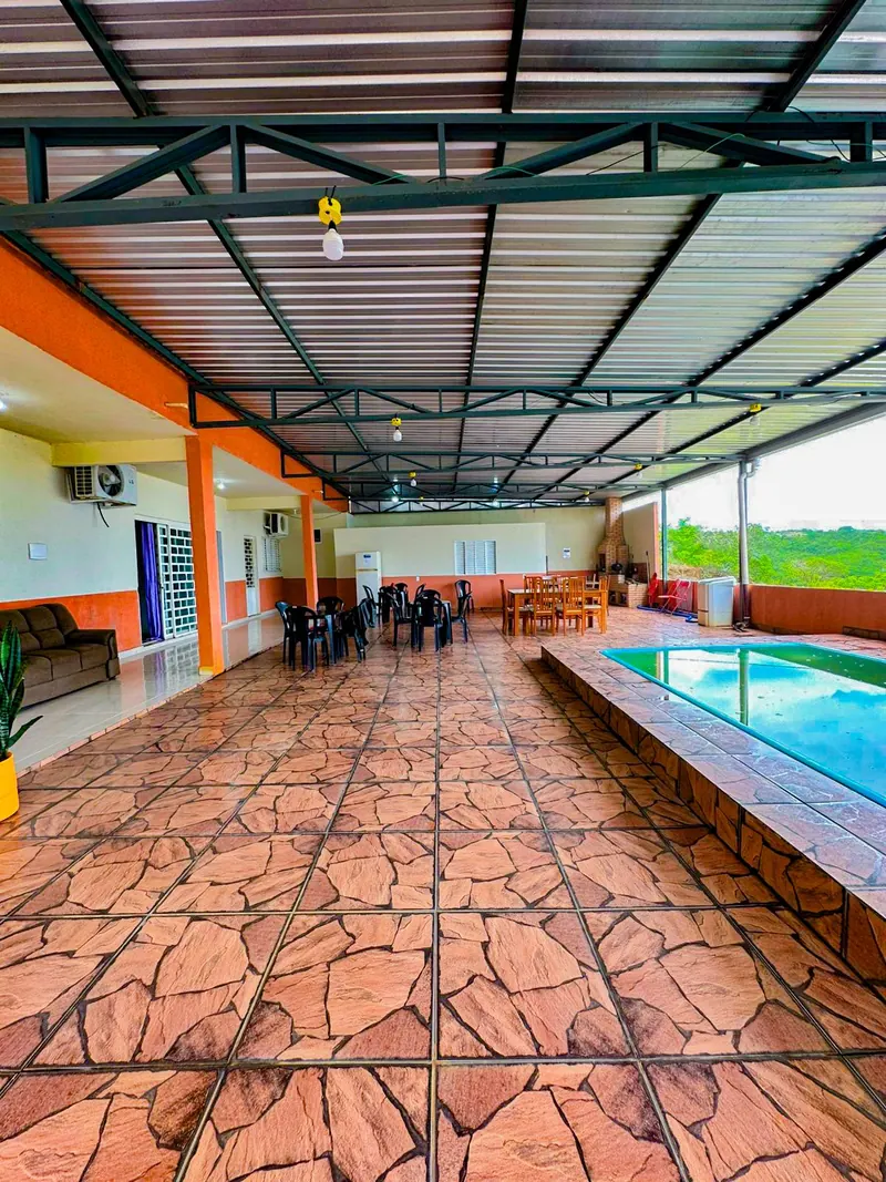 Salão coberto com piso de pedra e a piscina ao lado, com a mesa de madeira e as cadeiras ao fundo